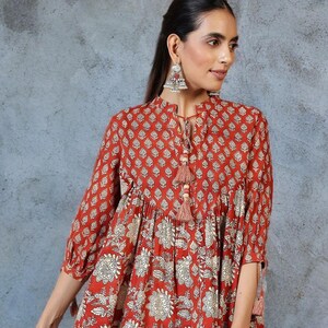 May include: A red tunic top with a floral and geometric pattern. The top has three-quarter sleeves and a mandarin collar with tassel detailing. The fabric is a warm red with cream-colored floral and geometric designs. The top is paired with white pants.