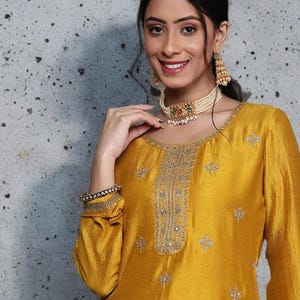May include: Mustard yellow silk kurta with silver embroidery on the neckline and scattered across the garment. The long-sleeved top is paired with a matching bracelet and a pearl choker necklace with gold and jewel accents. Gold earrings complete the look.