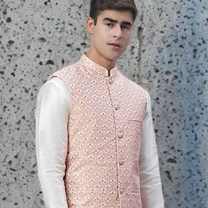 Op de afbeelding: Een perzikkleurig, patroonvest met een mandarijnkraag en gouden knopen. Het vest wordt gedragen over een wit shirt met lange mouwen. De outfit is formele kleding.