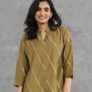 Puede incluir: Camisa verde oliva con un estampado de rayas diagonales blancas. La camisa tiene cuello mandarín y mangas tres cuartos. La modelo lleva la camisa.