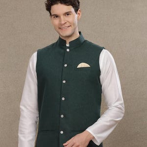 Op de afbeelding: Een donkergroen vest met een opstaande kraag en witte knopen. Het vest heeft een kleine zak met een goudkleurig accent. Het wordt gedragen over een wit shirt met lange mouwen.