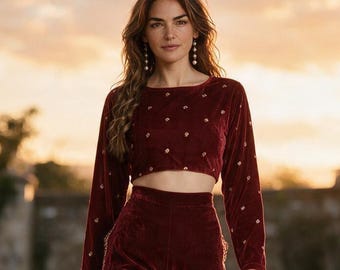 May include: A deep burgundy velvet two-piece set consisting of a long-sleeved cropped top and wide-leg trousers. The top features small embroidered floral accents, while the trousers have decorative side panels with intricate gold detailing.