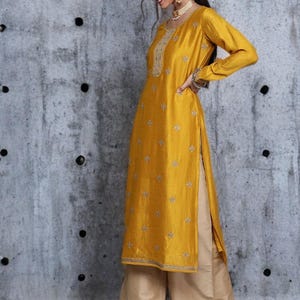 Puede incluir: Una t&uacute;nica de color amarillo mostaza de manga larga con bordados plateados en el escote y motivos florales dispersos por toda la prenda, combinada con pantalones beige de pierna ancha. La t&uacute;nica tiene aberturas laterales y un dobladillo recto.