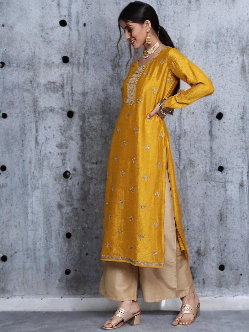 Puede incluir: Una t&uacute;nica de color amarillo mostaza de manga larga con bordados plateados en el escote y motivos florales dispersos por toda la prenda, combinada con pantalones beige de pierna ancha. La t&uacute;nica tiene aberturas laterales y un dobladillo recto.