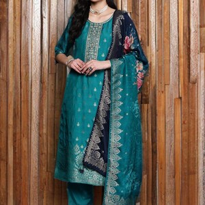 May include: Teal-colored salwar kameez ensemble with gold accents. The outfit includes a tunic, pants, and a shawl with floral and geometric patterns. The tunic features intricate embroidery around the neckline and bodice. The shawl has a navy blue border.