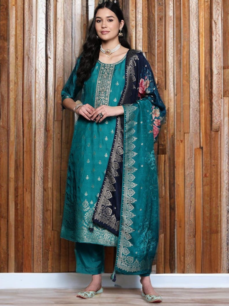 May include: Teal-colored salwar kameez ensemble with gold accents. The outfit includes a tunic, pants, and a shawl with floral and geometric patterns. The tunic features intricate embroidery around the neckline and bodice. The shawl has a navy blue border.