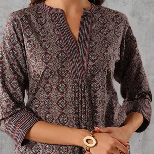 May include: A long-sleeved, burgundy tunic with a floral pattern. The tunic features a V-neck with decorative trim and three-quarter sleeves. A gold-toned bracelet with a large, circular design is visible on the wrist.