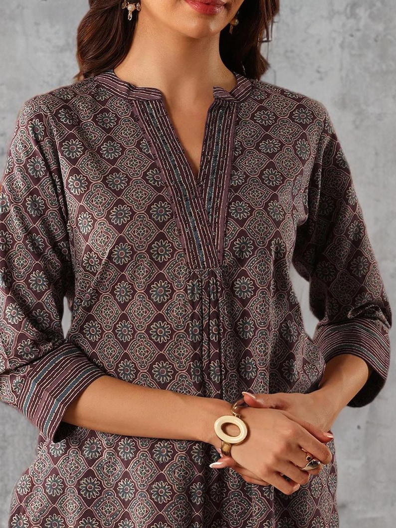 May include: A long-sleeved, burgundy tunic with a floral pattern. The tunic features a V-neck with decorative trim and three-quarter sleeves. A gold-toned bracelet with a large, circular design is visible on the wrist.