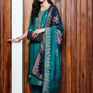 May include: Teal-colored salwar kameez ensemble with intricate embroidery on the neckline and sleeves. The outfit includes matching pants and a patterned shawl with floral accents. A black clutch and matching shoes complete the look.