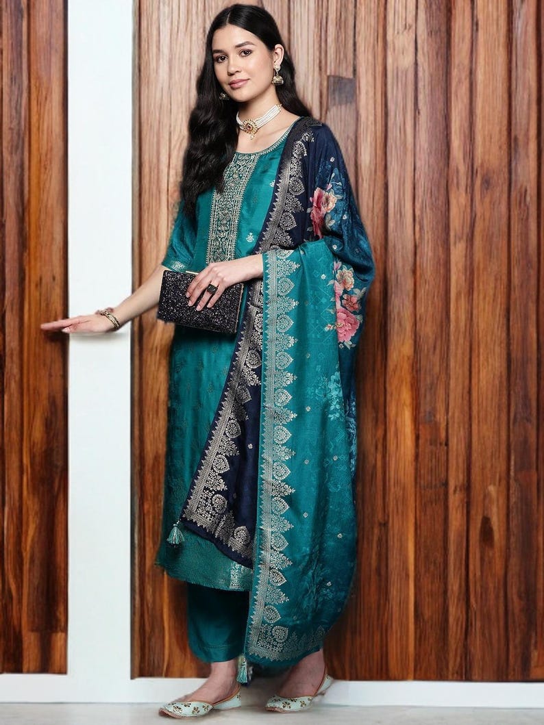 May include: Teal-colored salwar kameez ensemble with intricate embroidery on the neckline and sleeves. The outfit includes matching pants and a patterned shawl with floral accents. A black clutch and matching shoes complete the look.