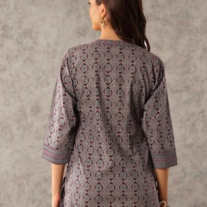 May include: A three-quarter sleeve tunic in a floral pattern. The tunic is primarily burgundy with a repeating floral design in white and blue. The sleeves and neckline have striped detailing. The model is wearing the tunic with blue jeans.
