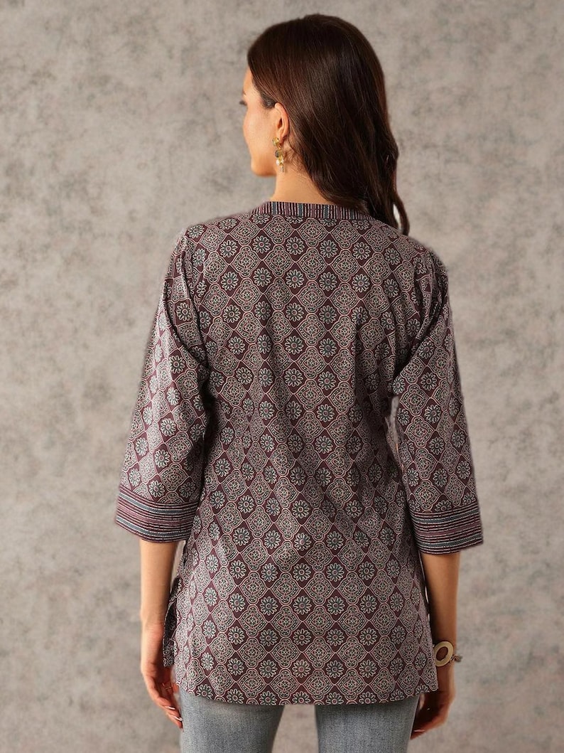 May include: A three-quarter sleeve tunic in a floral pattern. The tunic is primarily burgundy with a repeating floral design in white and blue. The sleeves and neckline have striped detailing. The model is wearing the tunic with blue jeans.