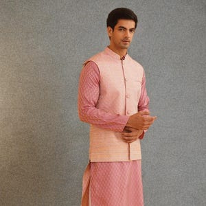 May include: A three-piece ensemble featuring a pink mandarin collar vest over a matching long-sleeved shirt and a knee-length tunic. The shirt and tunic have a textured pattern, and the vest has a lighter, textured fabric. The outfit is paired with cream-colored pants.
