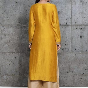 Puede incluir: Una t&uacute;nica de color amarillo mostaza de manga larga con un acabado ligeramente texturizado y bordados decorativos en los pu&ntilde;os, combinada con pantalones beige de pernera ancha. La t&uacute;nica llega por debajo de las caderas y el pantal&oacute;n hasta el suelo.