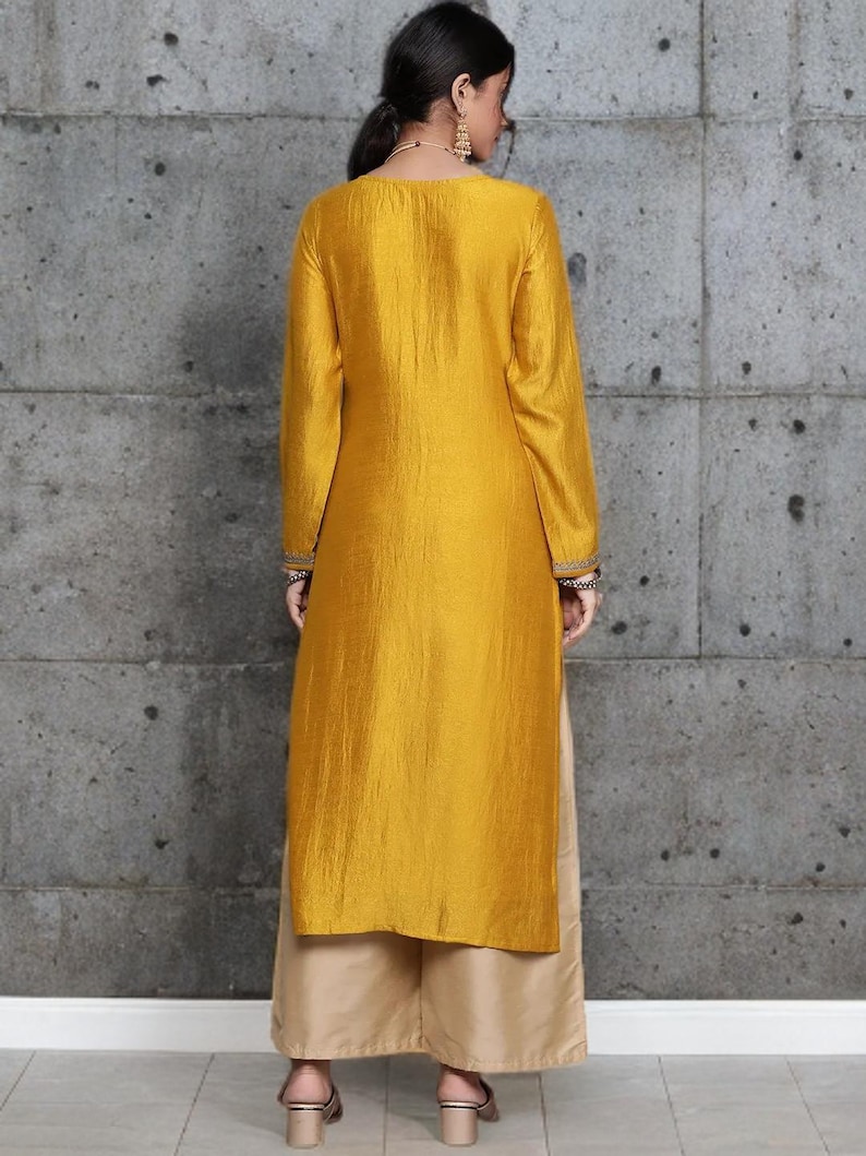 Puede incluir: Una t&uacute;nica de color amarillo mostaza de manga larga con un acabado ligeramente texturizado y bordados decorativos en los pu&ntilde;os, combinada con pantalones beige de pernera ancha. La t&uacute;nica llega por debajo de las caderas y el pantal&oacute;n hasta el suelo.