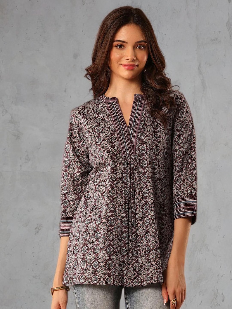 May include: A three-quarter sleeve tunic top with a V-neck. The top features a repeating floral pattern in shades of burgundy, gray, and white. The neckline and sleeve cuffs have striped detailing. The top is made of a soft, flowing fabric.
