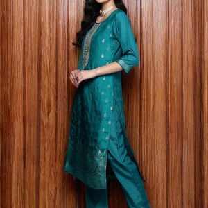 May include: Teal-colored Indian Kurta suit with intricate gold embroidery on the neckline and sleeves. The outfit includes a long tunic top and matching straight-leg pants. The model is wearing matching teal shoes.