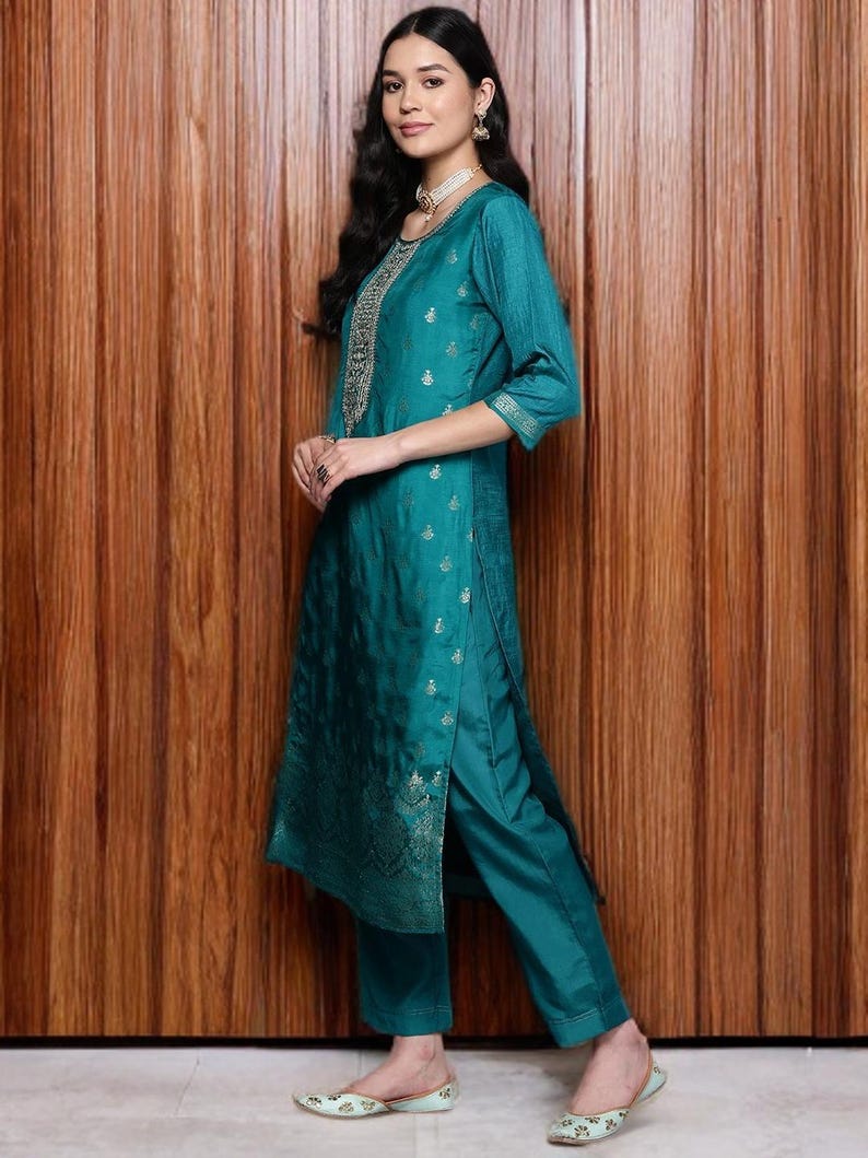May include: Teal-colored Indian Kurta suit with intricate gold embroidery on the neckline and sleeves. The outfit includes a long tunic top and matching straight-leg pants. The model is wearing matching teal shoes.