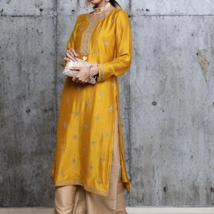 Puede incluir: Una t&uacute;nica de color amarillo mostaza con mangas largas, bordados plateados intrincados en el escote y motivos florales dispersos, combinada con pantalones beige de pierna ancha y un peque&ntilde;o bolso de mano rectangular adornado.