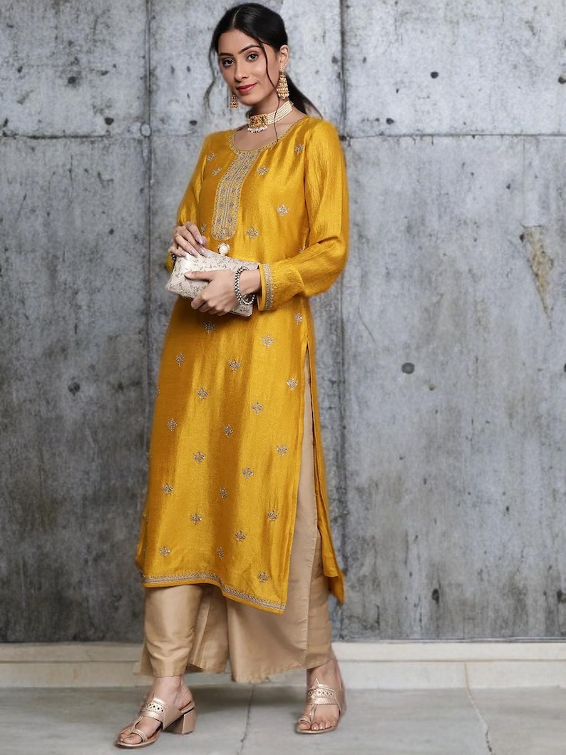 Puede incluir: Una t&uacute;nica de color amarillo mostaza con mangas largas, bordados plateados intrincados en el escote y motivos florales dispersos, combinada con pantalones beige de pierna ancha y un peque&ntilde;o bolso de mano rectangular adornado.