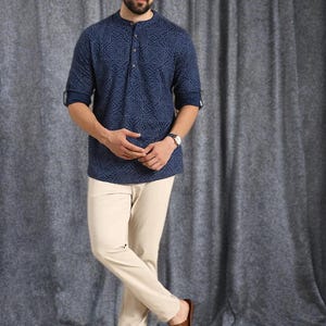 Pu&ograve; includere: Una camicia blu navy a maniche lunghe con un motivo geometrico, abbinata a pantaloni beige e sandali marroni. La camicia ha un colletto alla coreana e una chiusura con bottoni. I pantaloni sono a gamba dritta. I sandali sono in pelle.