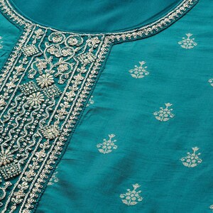 May include: Teal-colored garment with intricate embroidery around the neckline and down the front. The embroidery is a mix of floral and geometric patterns in a light, contrasting color. The fabric has a subtle sheen and small, repeating floral motifs.