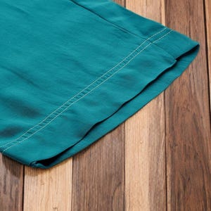 May include: Close-up of teal fabric with a white stitched seam. The fabric is folded over, revealing the underside. The background is a wooden surface with vertical planks in varying shades of brown.