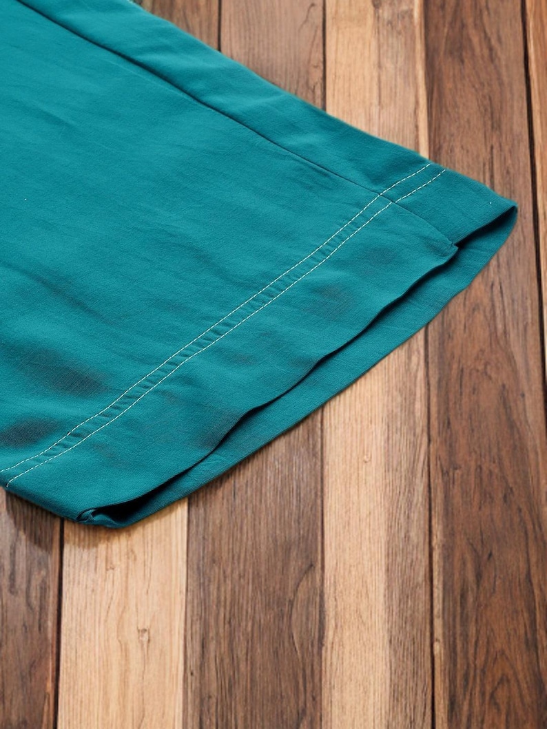 May include: Close-up of teal fabric with a white stitched seam. The fabric is folded over, revealing the underside. The background is a wooden surface with vertical planks in varying shades of brown.