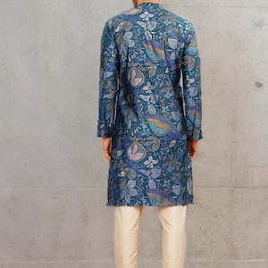May include: A teal long-sleeved tunic with a floral pattern, paired with cream-colored pants. The tunic features a mandarin collar and a button-down front. The floral design includes shades of blue, purple, and gold. The pants are a straight-leg style.