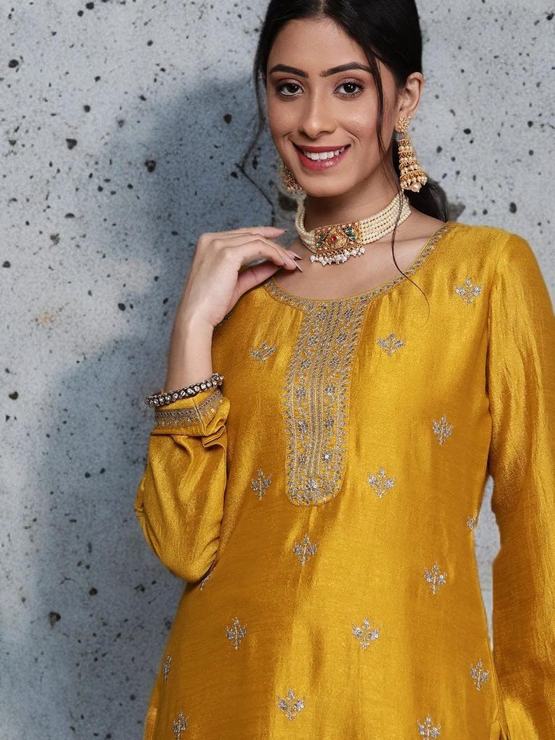 Puede incluir: Una t&uacute;nica de color amarillo mostaza con mangas largas, motivos florales bordados en plata, un panel central vertical con detalles plateados, un collar de perlas de varias hileras y pendientes dorados.