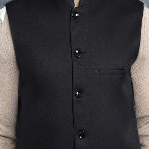 Op de afbeelding: Een zwarte gilet met een opstaande kraag en vier zwarte knopen. De gilet heeft een klein zakje aan de linkerkant. De persoon draagt een wit overhemd en een beige trui eronder.