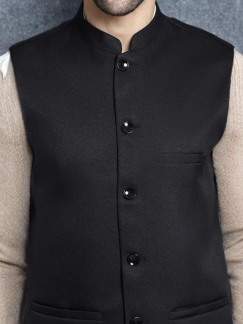 Op de afbeelding: Een zwarte gilet met een opstaande kraag en vier zwarte knopen. De gilet heeft een klein zakje aan de linkerkant. De persoon draagt een wit overhemd en een beige trui eronder.