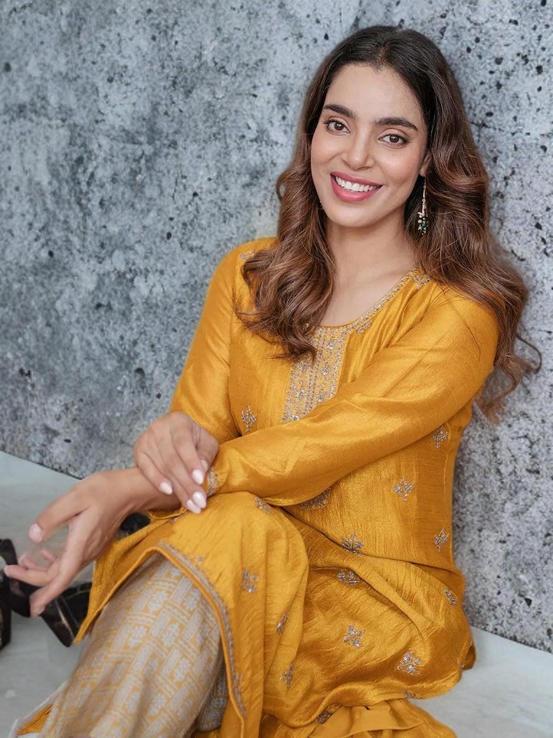 Puede incluir: Una t&uacute;nica de color amarillo mostaza de manga larga con delicados bordados plateados a lo largo del escote y motivos florales dispersos por la tela, combinada con pantalones de color beige claro con un sutil patr&oacute;n geom&eacute;trico.