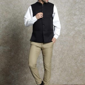 Op de afbeelding: Een zwart vest met een opstaande kraag en knoopsluiting, gedragen over een wit overhemd. De outfit omvat beige broeken en donkerbruine leren schoenen. De ensemble is een klassieke, formele stijl.