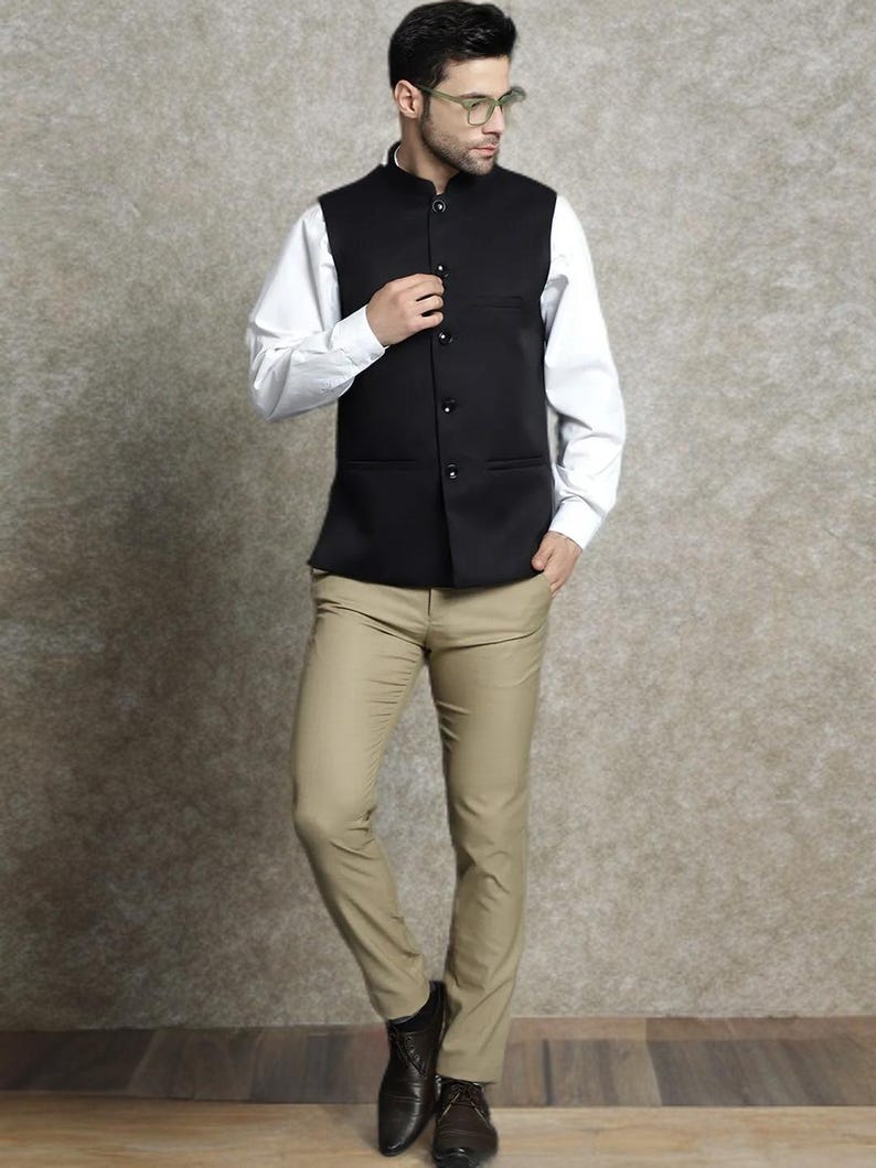Op de afbeelding: Een zwart vest met een opstaande kraag en knoopsluiting, gedragen over een wit overhemd. De outfit omvat beige broeken en donkerbruine leren schoenen. De ensemble is een klassieke, formele stijl.