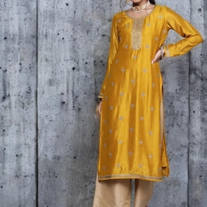 Puede incluir: Una t&uacute;nica de color amarillo mostaza de manga larga con motivos florales bordados en plata, cuello redondo con un panel plateado decorativo y aberturas laterales, combinada con pantalones beige de pierna ancha y sandalias.