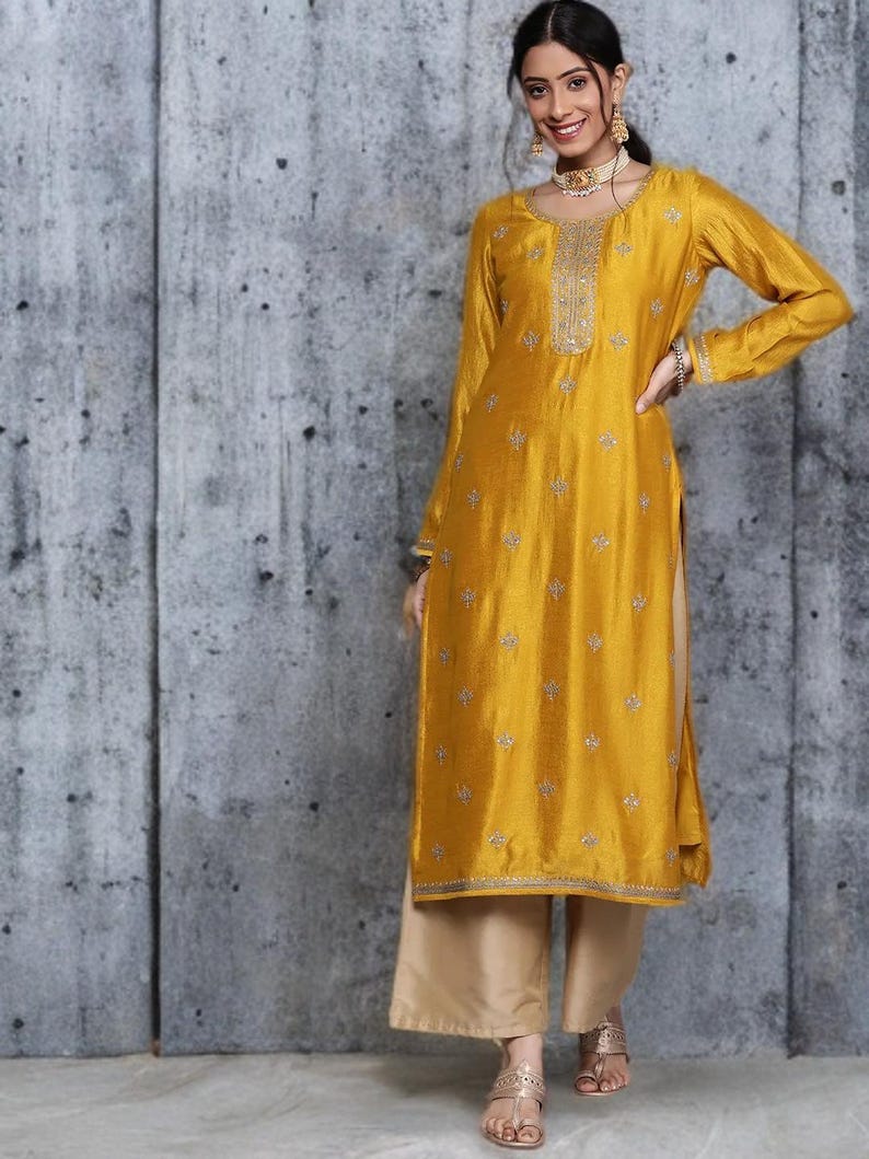 Puede incluir: Una t&uacute;nica de color amarillo mostaza de manga larga con motivos florales bordados en plata, cuello redondo con un panel plateado decorativo y aberturas laterales, combinada con pantalones beige de pierna ancha y sandalias.