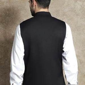 Op de afbeelding: Een zwart vest met een hoge kraag, gedragen over een wit shirt met lange mouwen. Het vest bedekt de voor- en achterkant van de torso, met de witte mouwen van het shirt zichtbaar. De outfit staat tegen een neutrale achtergrond.