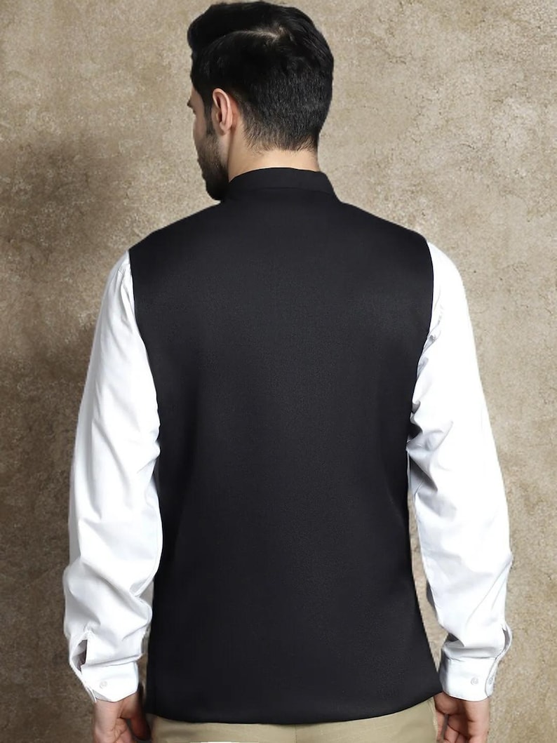 Op de afbeelding: Een zwart vest met een hoge kraag, gedragen over een wit shirt met lange mouwen. Het vest bedekt de voor- en achterkant van de torso, met de witte mouwen van het shirt zichtbaar. De outfit staat tegen een neutrale achtergrond.
