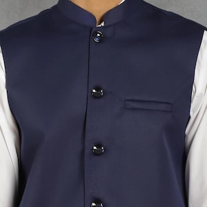 Op de afbeelding: Een marineblauw vest met een opstaande kraag en drie zwarte knopen. Het vest heeft een kleine borstzak en wordt over een wit overhemd gedragen. Een formeel kledingstuk.