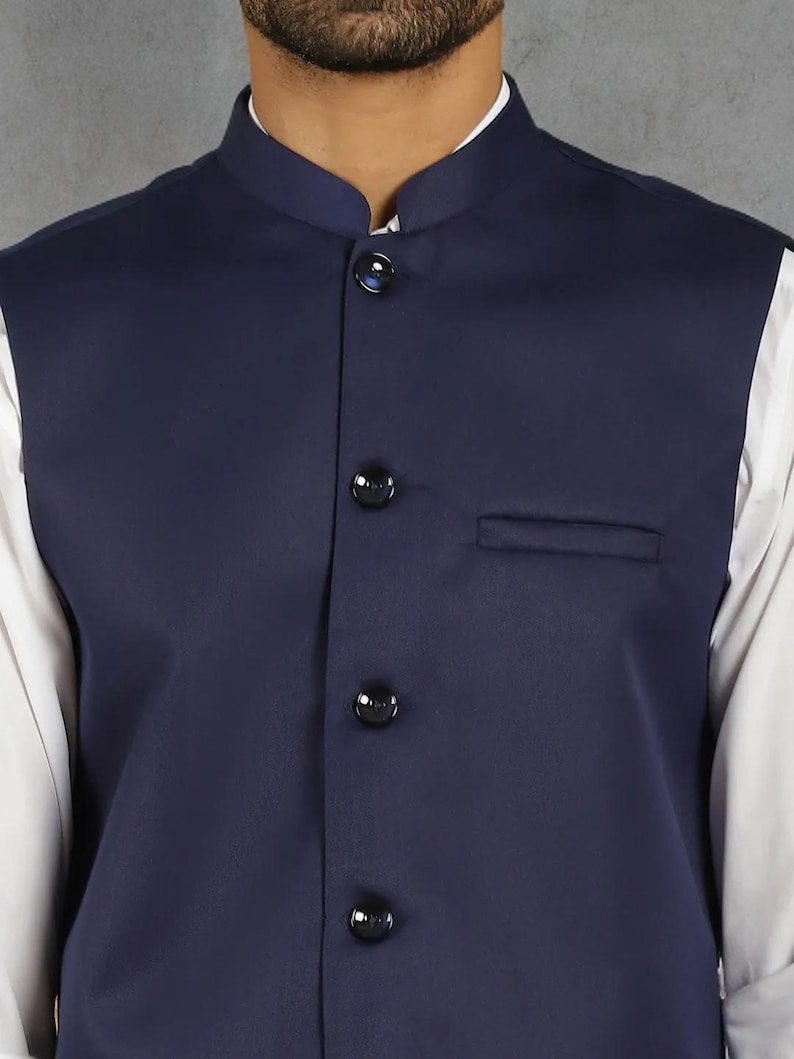 Op de afbeelding: Een marineblauw vest met een opstaande kraag en drie zwarte knopen. Het vest heeft een kleine borstzak en wordt over een wit overhemd gedragen. Een formeel kledingstuk.