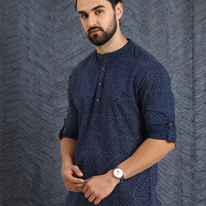 Pu&ograve; includere: Una camicia blu navy con motivo geometrico e colletto alla coreana. La camicia ha maniche lunghe arrotolate sui gomiti. &Egrave; visibile un orologio da polso con cinturino blu. La camicia &egrave; abbinata a pantaloni beige.