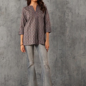 May include: A woman wearing a patterned, long-sleeve tunic and light-wash flared jeans. The tunic is a muted purple with a repeating geometric design. The jeans have a classic five-pocket design. The woman is also wearing gold-colored sandals.