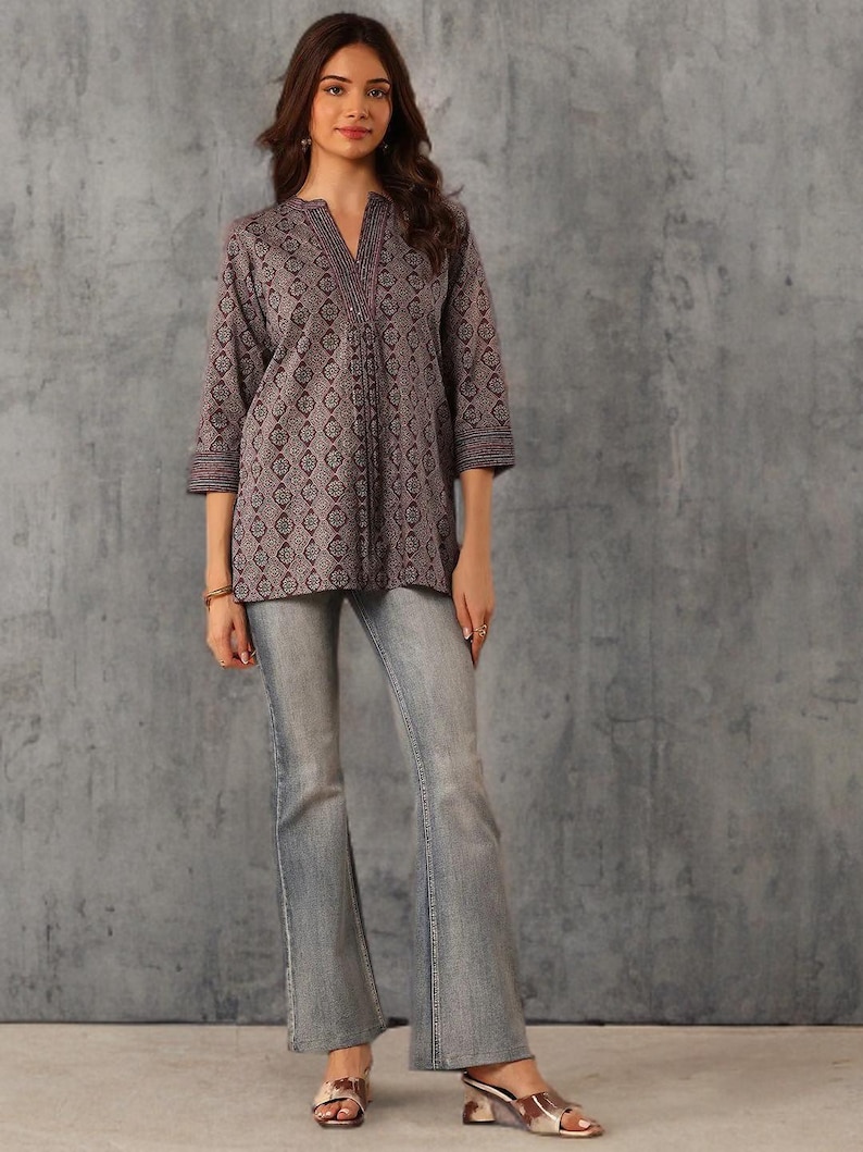 May include: A woman wearing a patterned, long-sleeve tunic and light-wash flared jeans. The tunic is a muted purple with a repeating geometric design. The jeans have a classic five-pocket design. The woman is also wearing gold-colored sandals.