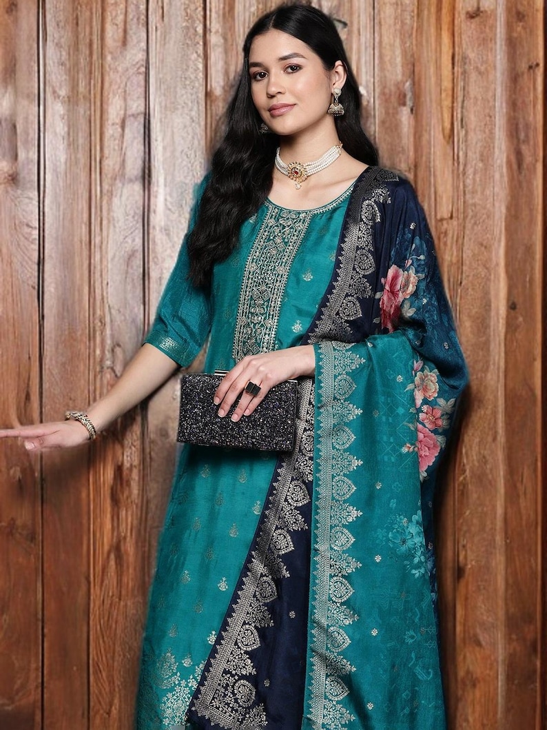 May include: A teal-colored formal dress with gold embroidery and a matching shawl featuring floral patterns. The dress is paired with a black beaded clutch and jewelry. The outfit is set against a wooden background.
