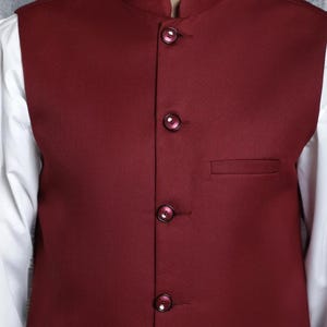 Op de afbeelding: Een bordeauxrode gilet met een opstaande kraag en vier bijpassende knopen. De gilet heeft een klein zakje aan de linkerkant en wordt over een wit overhemd gedragen. De gilet is effen van kleur en lijkt van een gladde stof te zijn.