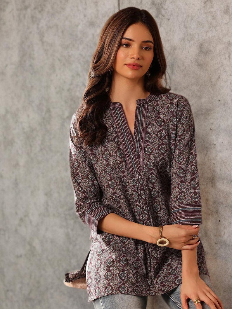 May include: A dark purple tunic with a geometric floral pattern. The tunic has a V-neckline with vertical striped detailing and three-quarter sleeves. The fabric appears to be a soft, woven material.