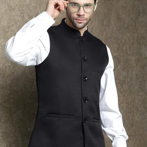 Op de afbeelding: Een zwart vest met een opstaande kraag en knoopsluiting, gedragen over een wit overhemd. Het vest heeft twee voorzakken. De persoon op de foto draagt een bril met groen montuur.
