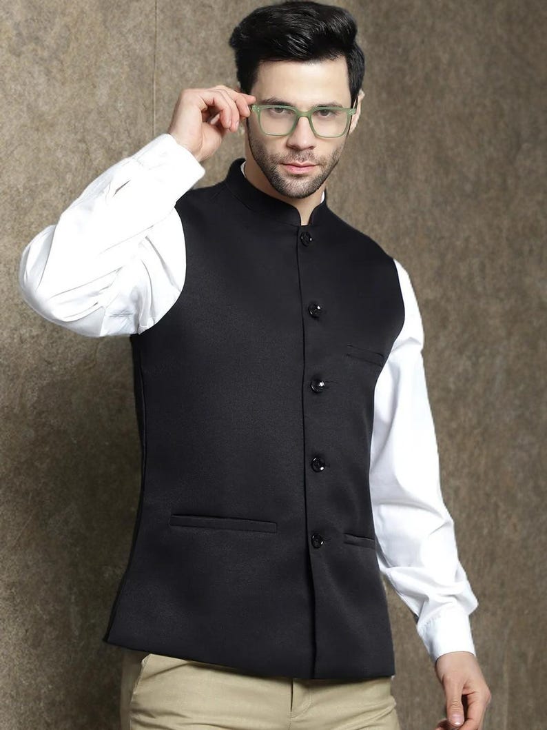 Op de afbeelding: Een zwart vest met een opstaande kraag en knoopsluiting, gedragen over een wit overhemd. Het vest heeft twee voorzakken. De persoon op de foto draagt een bril met groen montuur.