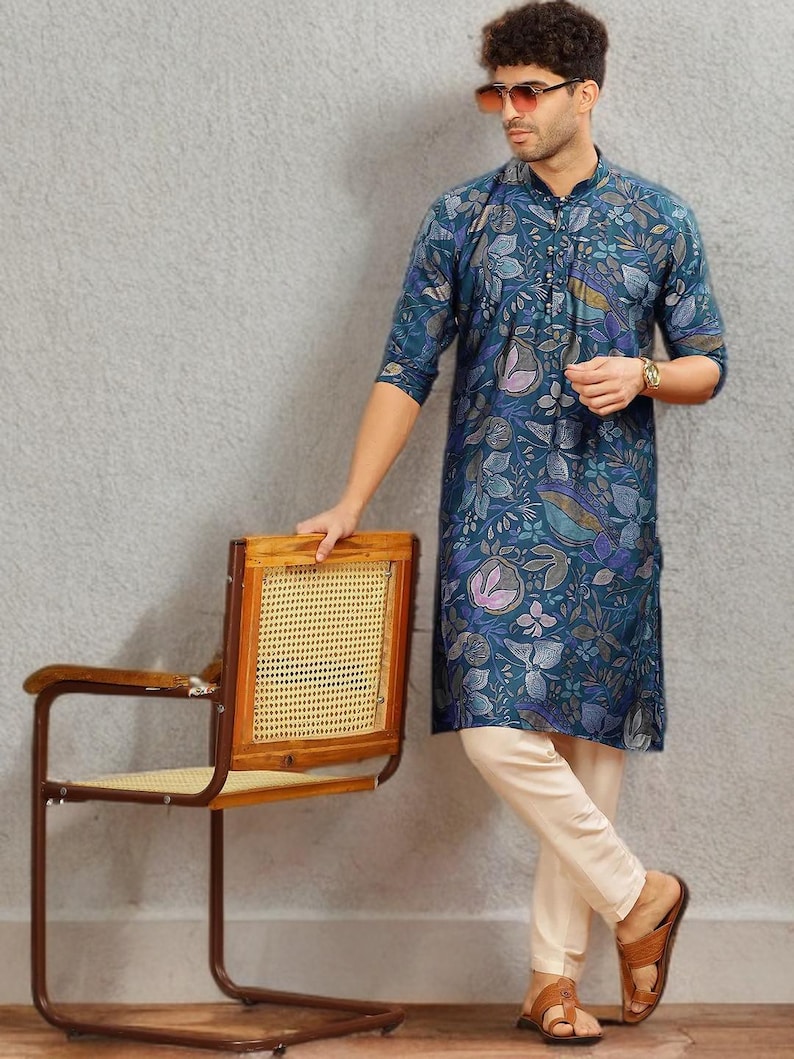 May include: A teal floral print kurta with cream-colored pants. The kurta features a button-down front and a mandarin collar. The outfit is paired with brown leather sandals. A wooden chair with a woven back is also in the image.